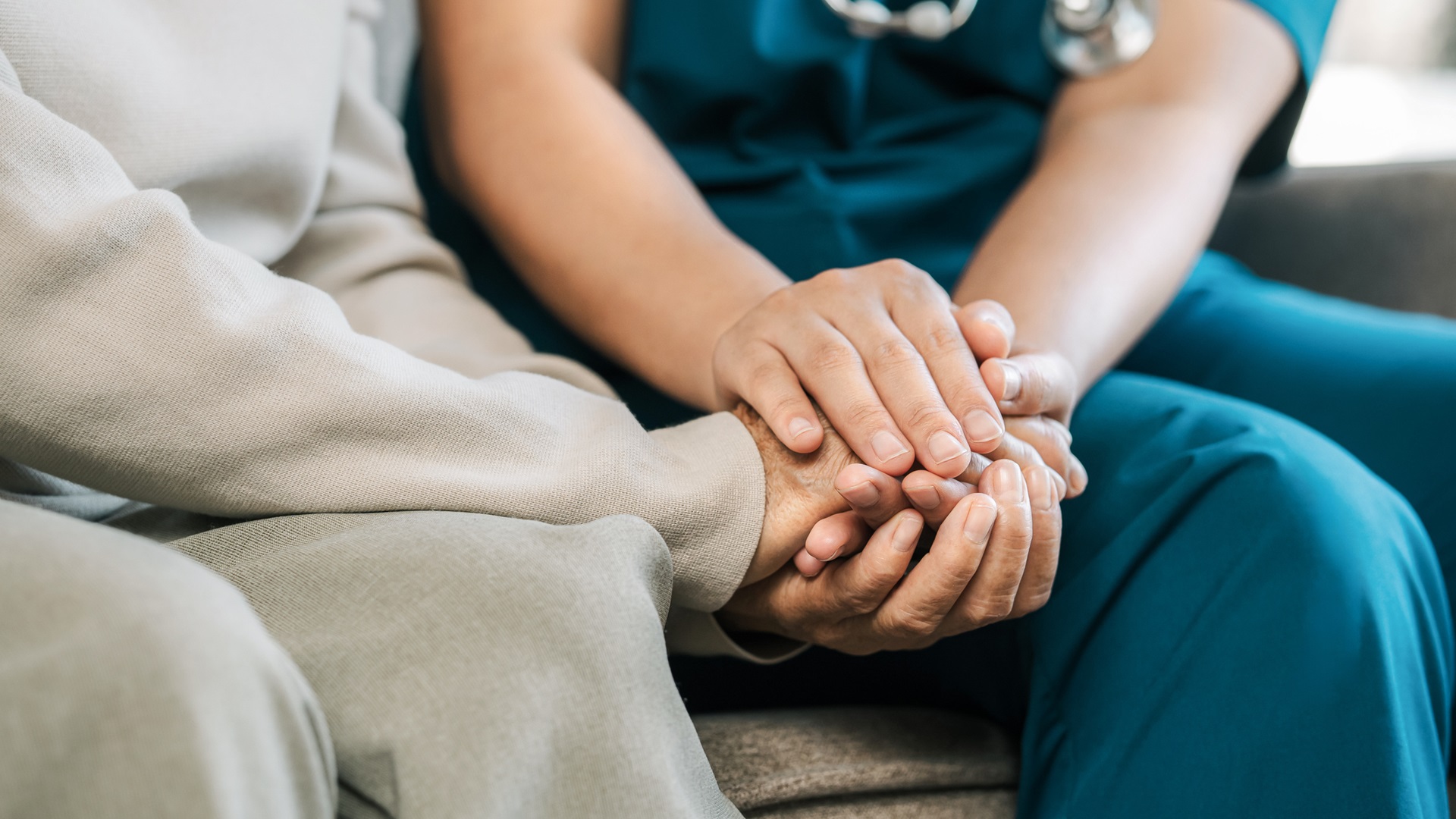 Ruce pečovatelky drží ruce starší osoby, symbolizující péči a podporu. Zdůrazňuje domácí zdravotní péči.