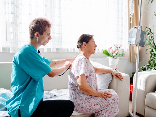 Zdravotník poslouchá stetoskopem záda starší ženy, která sedí na pohovce. Ilustruje domácí zdravotní péči.