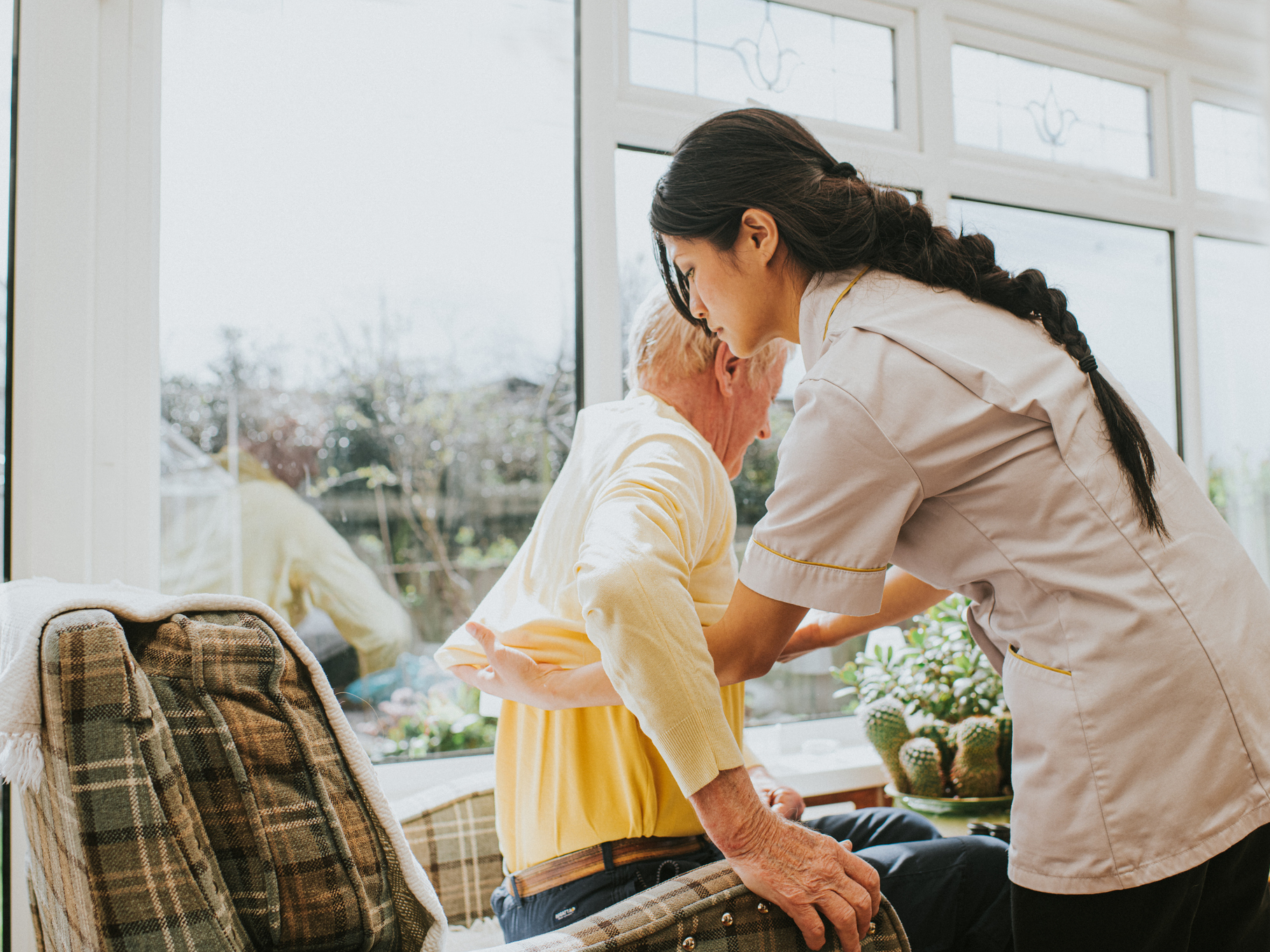 Pečovatelka pomáhá staršímu muži vstát ze židle u okna. Zobrazuje podporu a péči v domácím prostředí.