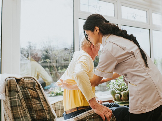 Pečovatelka pomáhá staršímu muži vstát ze židle u okna. Zobrazuje podporu a péči v domácím prostředí.