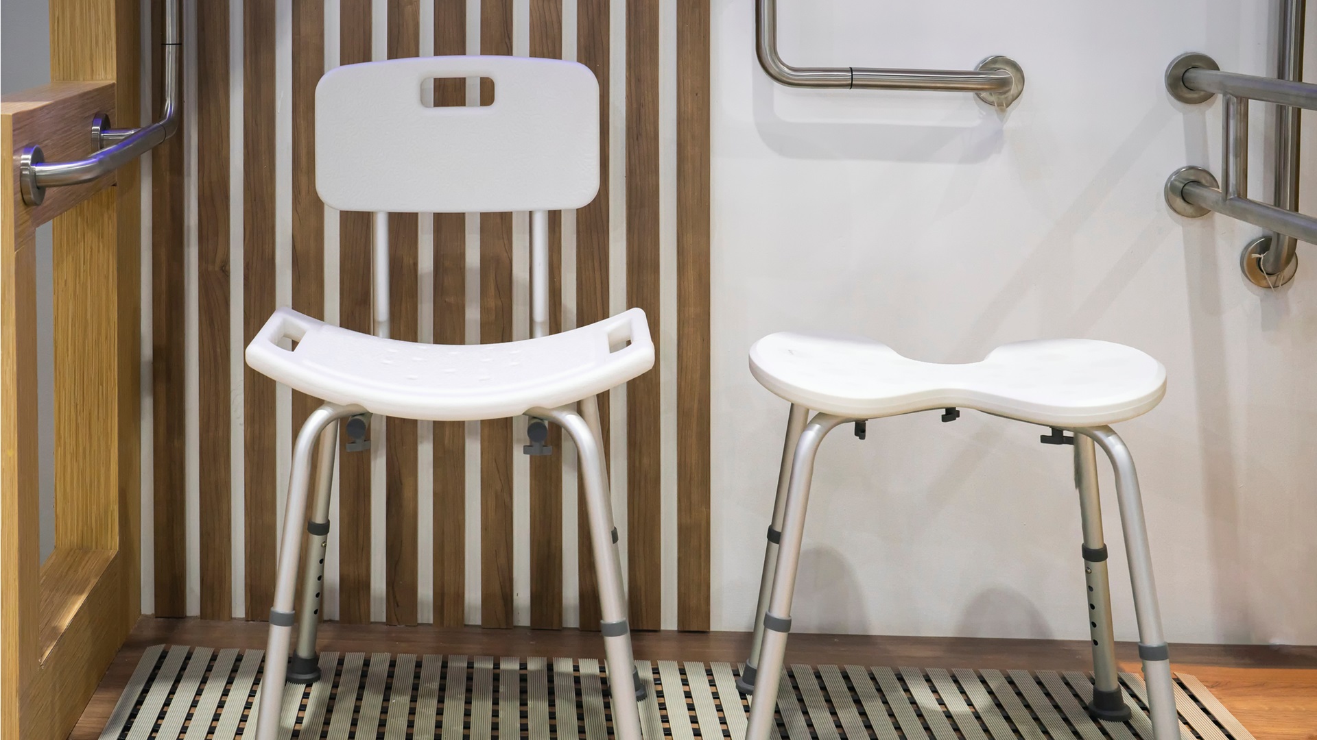 Dvě bílé sprchové židle a madla na stěně v koupelně. Zdůrazňuje pomůcky pro hygienu osob s omezenou hybností.