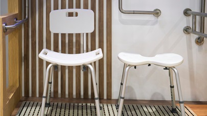 Dvě bílé sprchové židle a madla na stěně v koupelně. Zdůrazňuje pomůcky pro hygienu osob s omezenou hybností.
