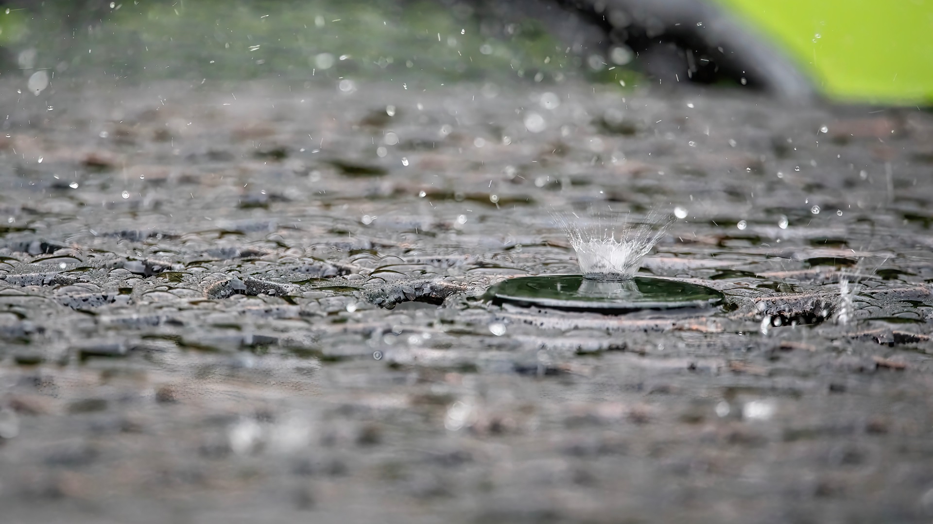Kapky deště dopadají na mokrý, tmavý povrch, vytvářejíce malé stříkance. V popředí je vidět jeden výrazný stříkanec.
