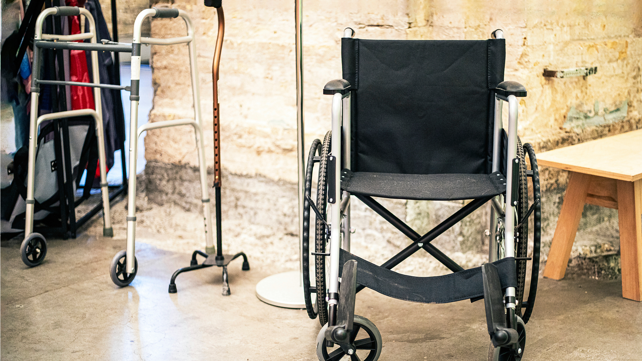 Invalidní vozík, chodítka a berle stojí v místnosti, připravené k použití. Symbolizují pomůcky pro usnadnění pohybu a péče.