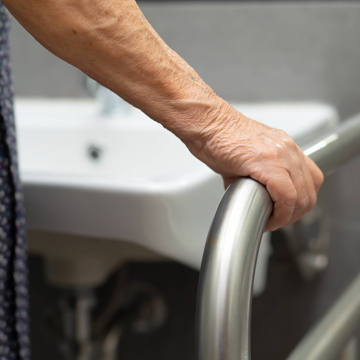 Starší osoba se drží madla v koupelně, s umyvadlem v pozadí. Zdůrazňuje bezpečnost a přístupnost v domácnosti.