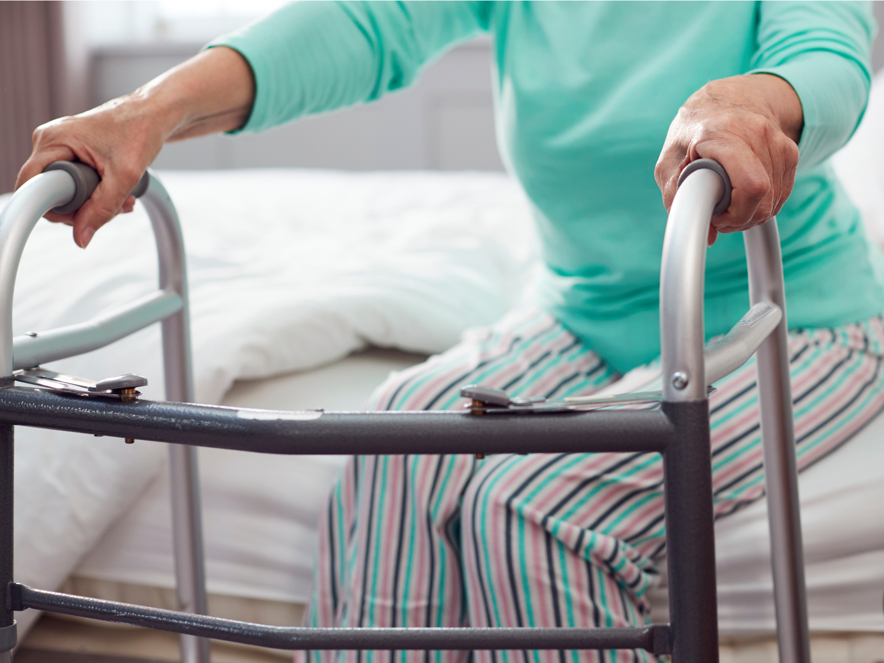 Starší žena v pyžamu sedí na posteli a drží se chodítka, připravena vstát.