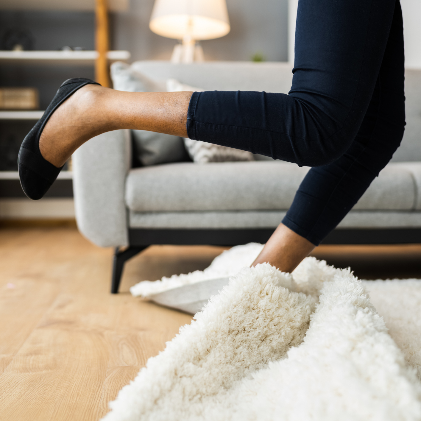 Noha zakopávající o zvednutý koberec v obývacím pokoji. Upozorňuje na rizika pádu v domácnosti.