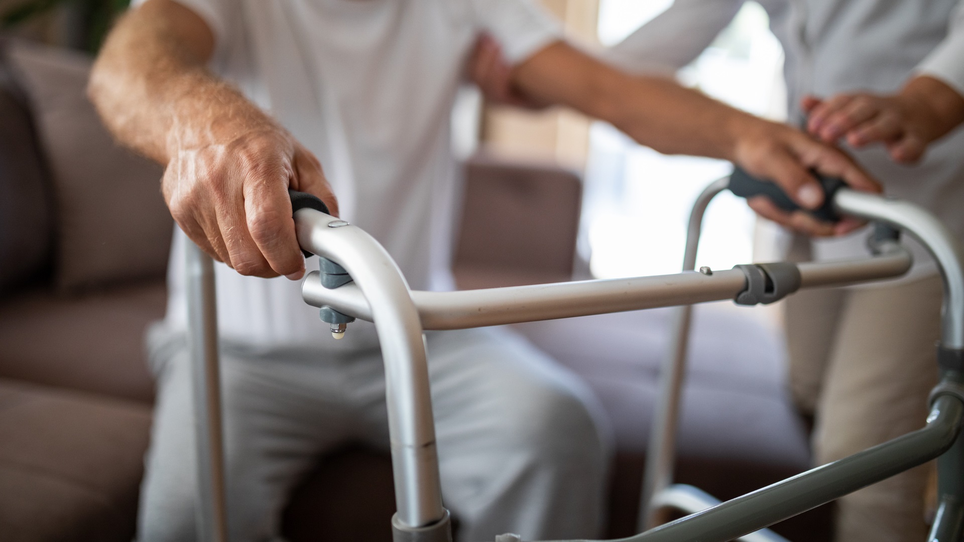 Starší muž se drží chodítka, zatímco mu druhá osoba pomáhá.