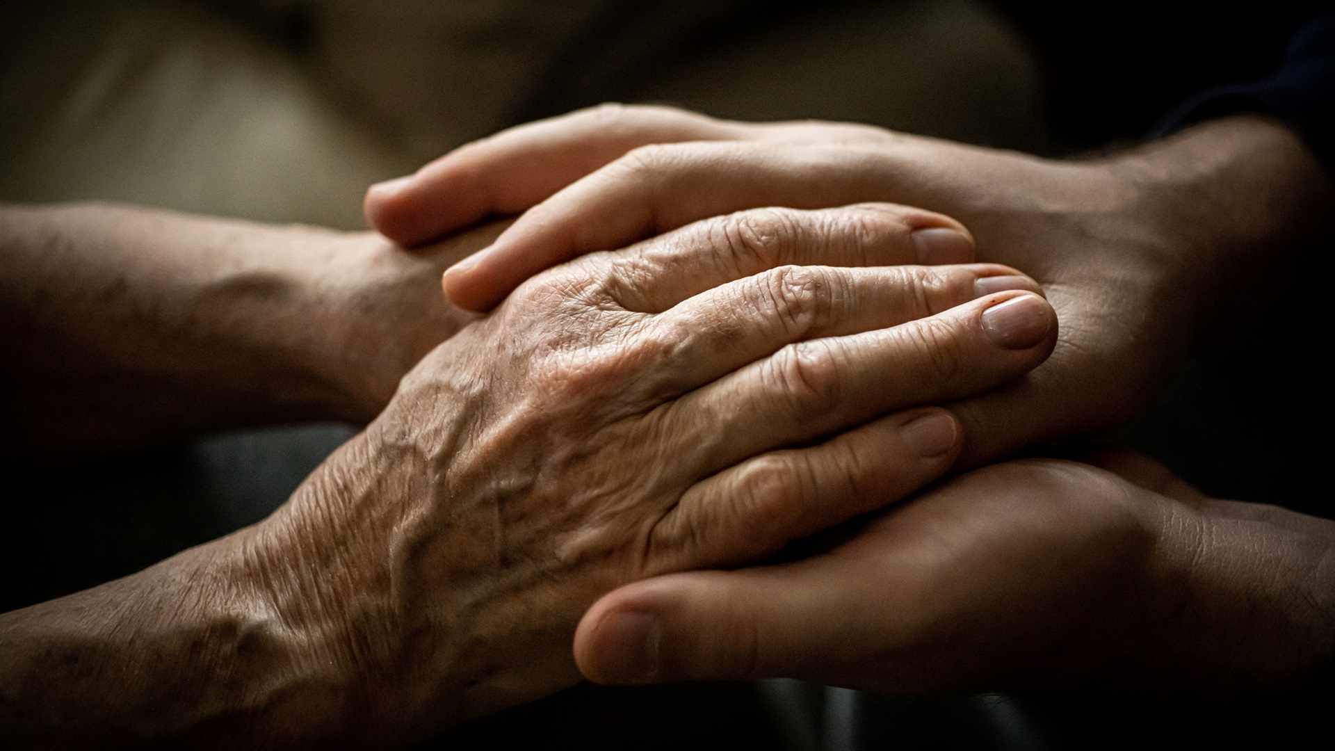 Detailní záběr na ruce staršího člověka, které drží a utěšuje mladší člověk. Ukazuje se podpora a péče.