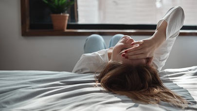 Žena leží na posteli s rukou přes oči, což naznačuje únavu nebo vyčerpání. Symbolizuje syndrom vyhoření.