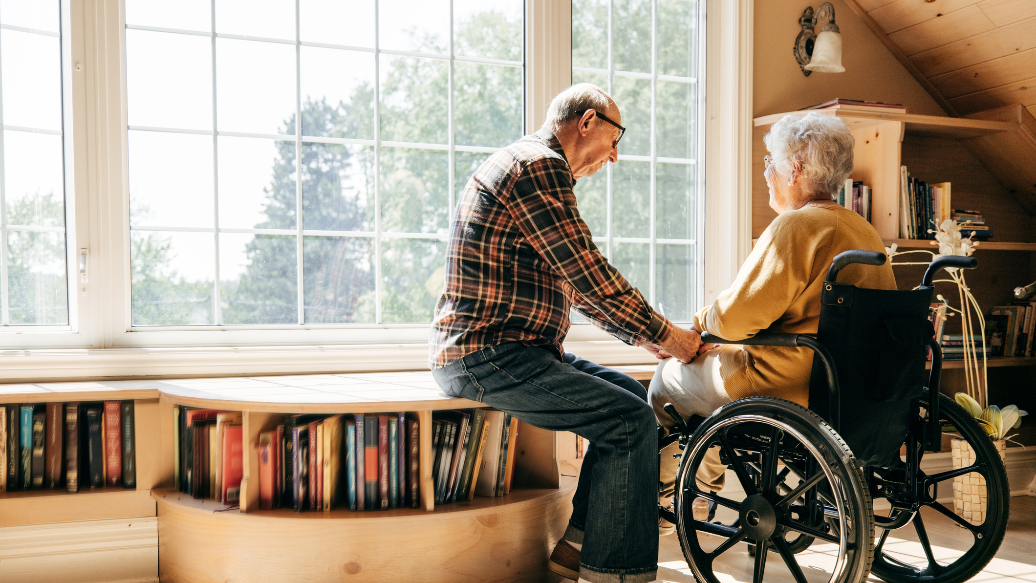 Starší muž sedí u okna a drží za ruku ženu na invalidním vozíku. Vyzařuje z nich klid a vzájemná podpora.